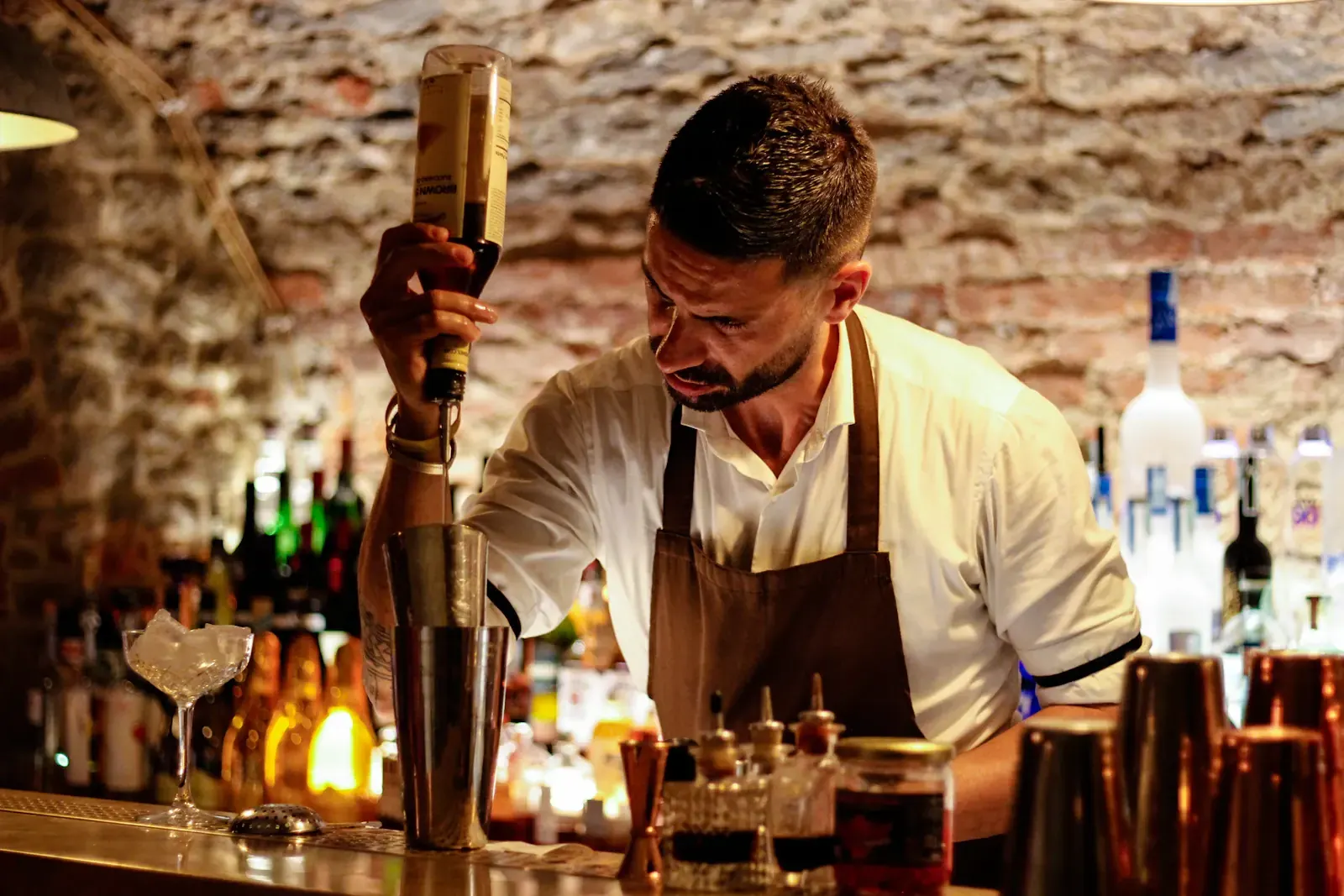 A bartender in an apron mixes a drink behind a bar